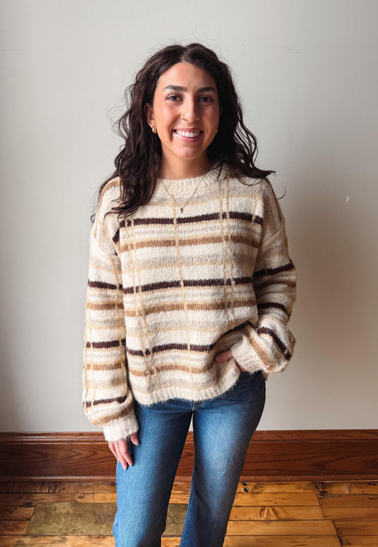A woman with long dark hair stands indoors wearing a fuzzy knit sweater with horizontal beige, brown, and cream stripes, paired with blue jeans, against a plain light gray wall and wooden floor.