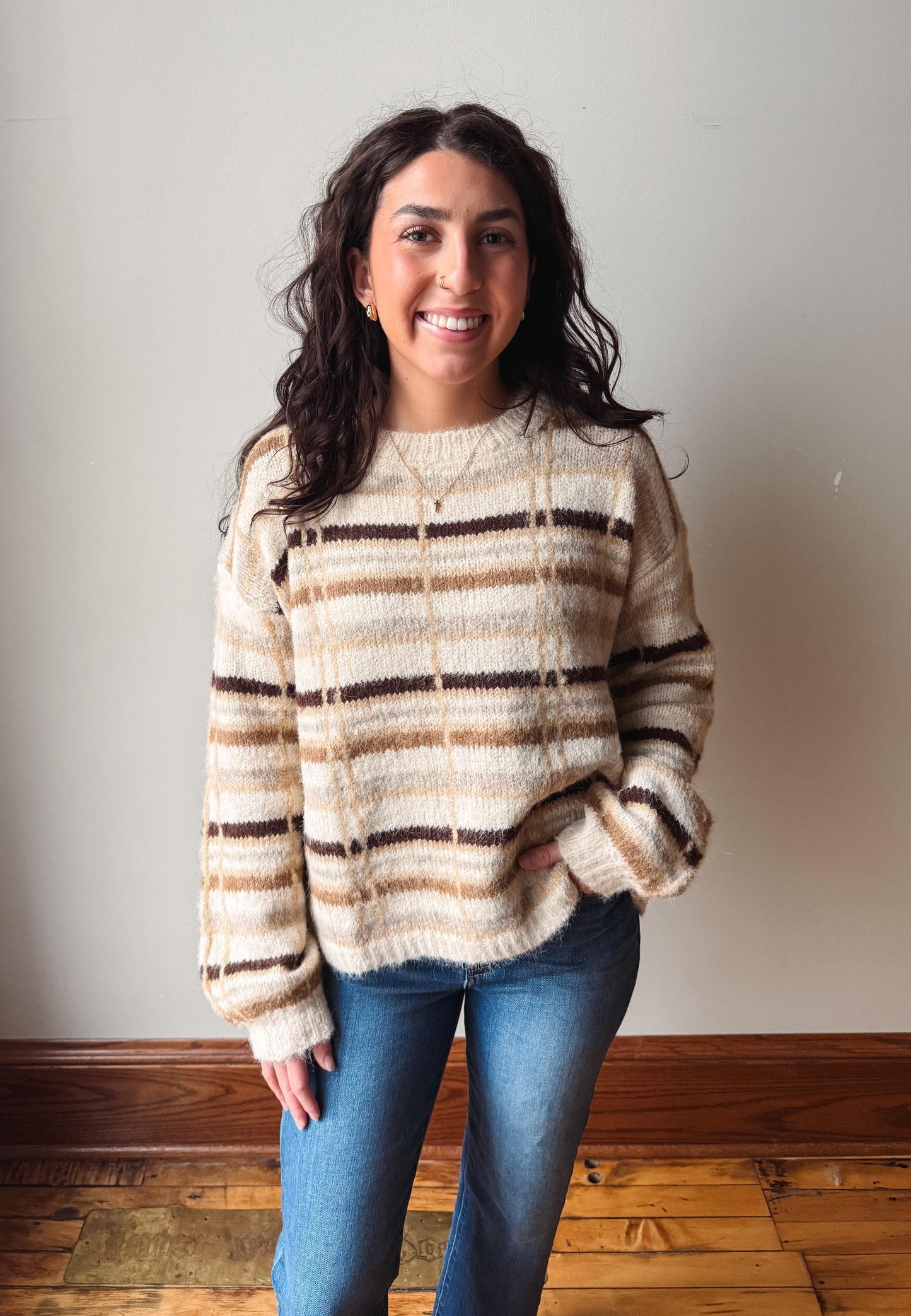 A woman with long dark hair stands indoors wearing a fuzzy knit sweater with horizontal beige, brown, and cream stripes, paired with blue jeans, against a plain light gray wall and wooden floor.