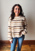 A woman with long dark hair stands indoors wearing a fuzzy knit sweater with horizontal beige, brown, and cream stripes, paired with blue jeans, against a plain light gray wall and wooden floor.