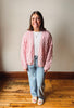 A woman stands in a room with a beige wall and wooden floor, wearing a pink cable-knit cardigan over a white top and light blue jeans.