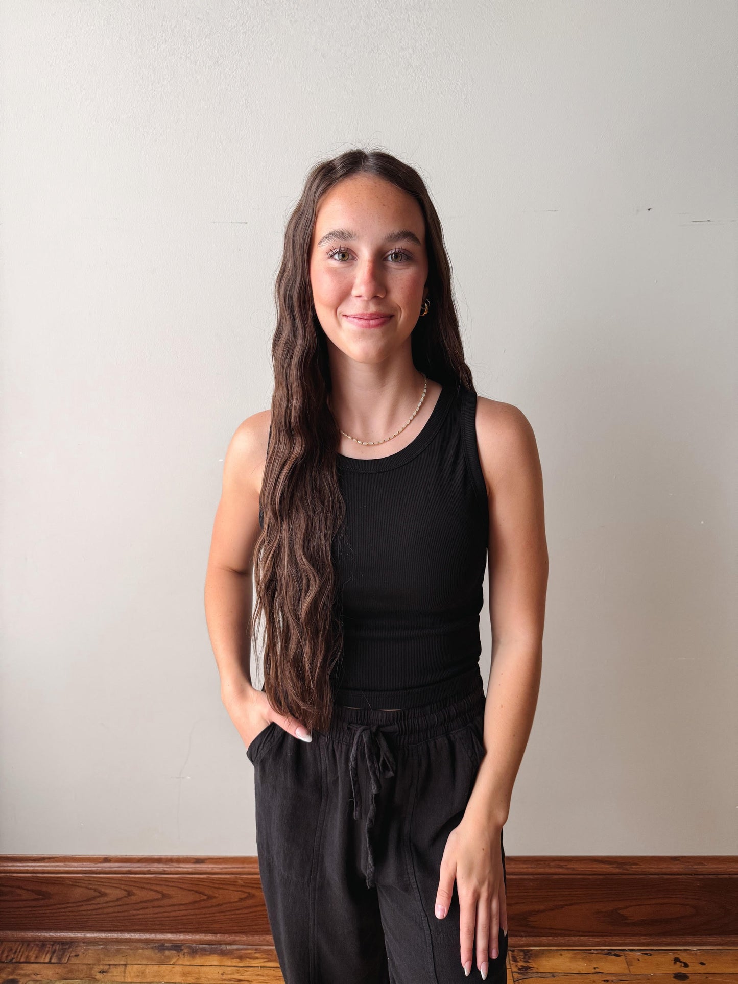 Woman wearing a black sleeveless top and black pants against a plain wall.