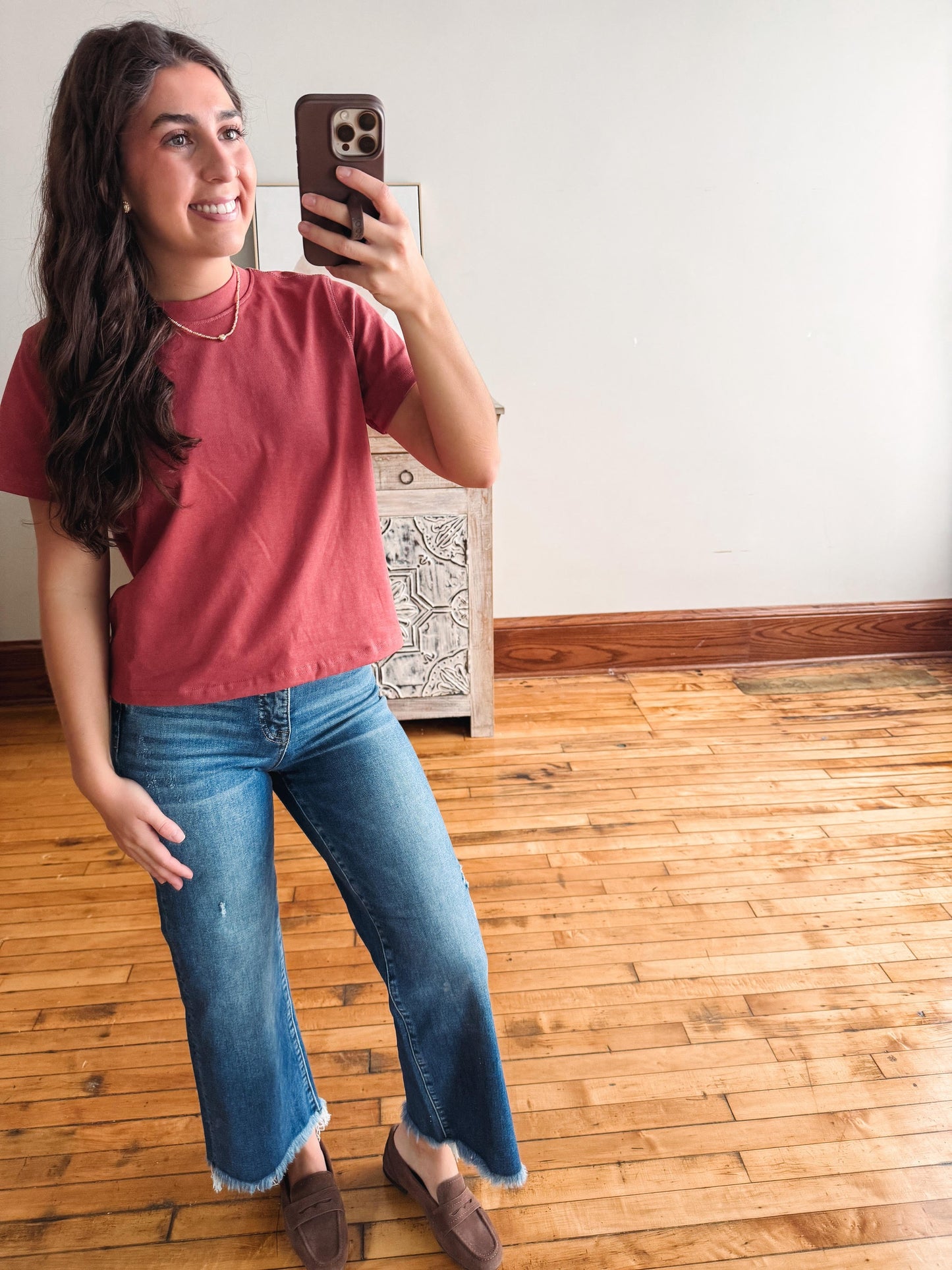 A woman taking a mirror selfie in a room with hardwood floors and a white wall, wearing a dusty rose short-sleeve t-shirt, blue wide-leg jeans, and brown leather loafers.