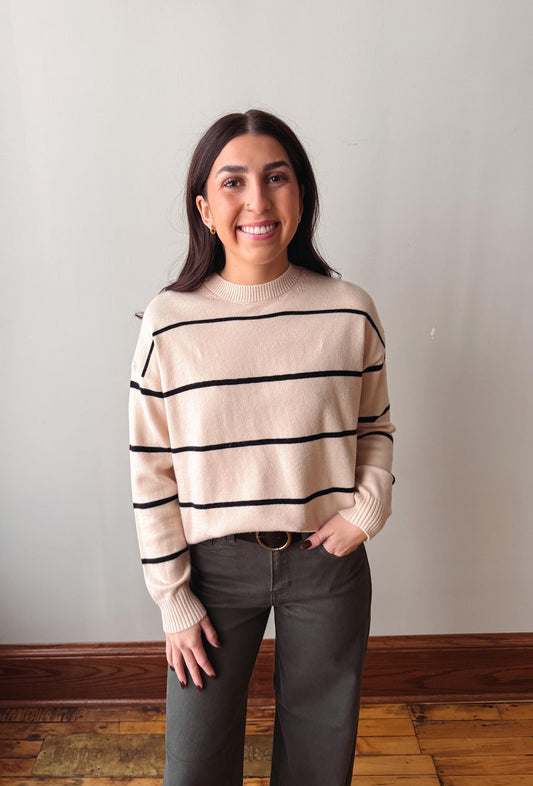 A woman smiling and posing in a cream-colored knit sweater with black horizontal stripes, paired with dark gray pants, standing against a plain light gray wall.
