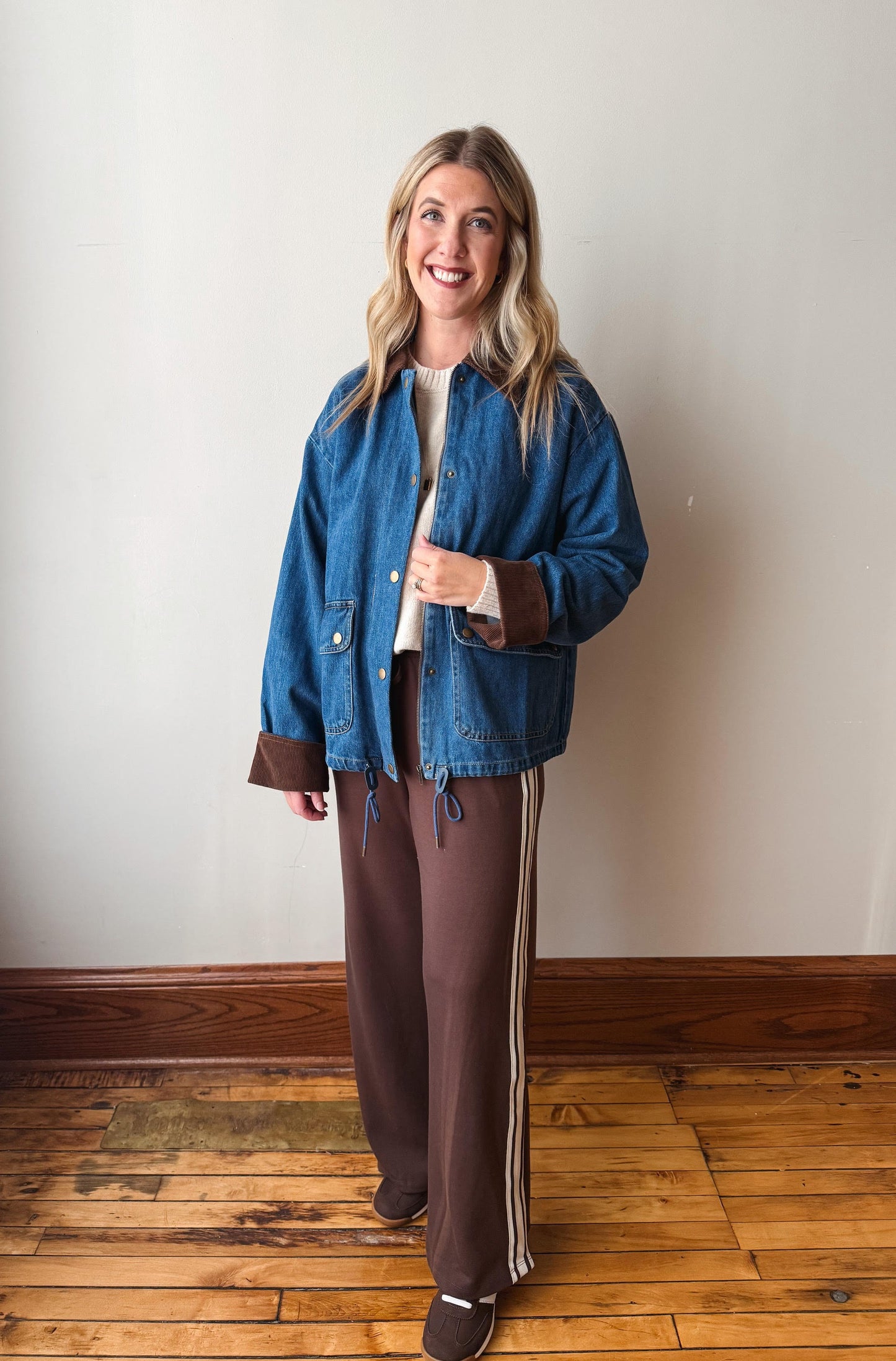 A woman stands in a room with a beige wall and wooden floor, wearing a blue denim jacket with brown trim over a white top and brown pants with white side stripes.