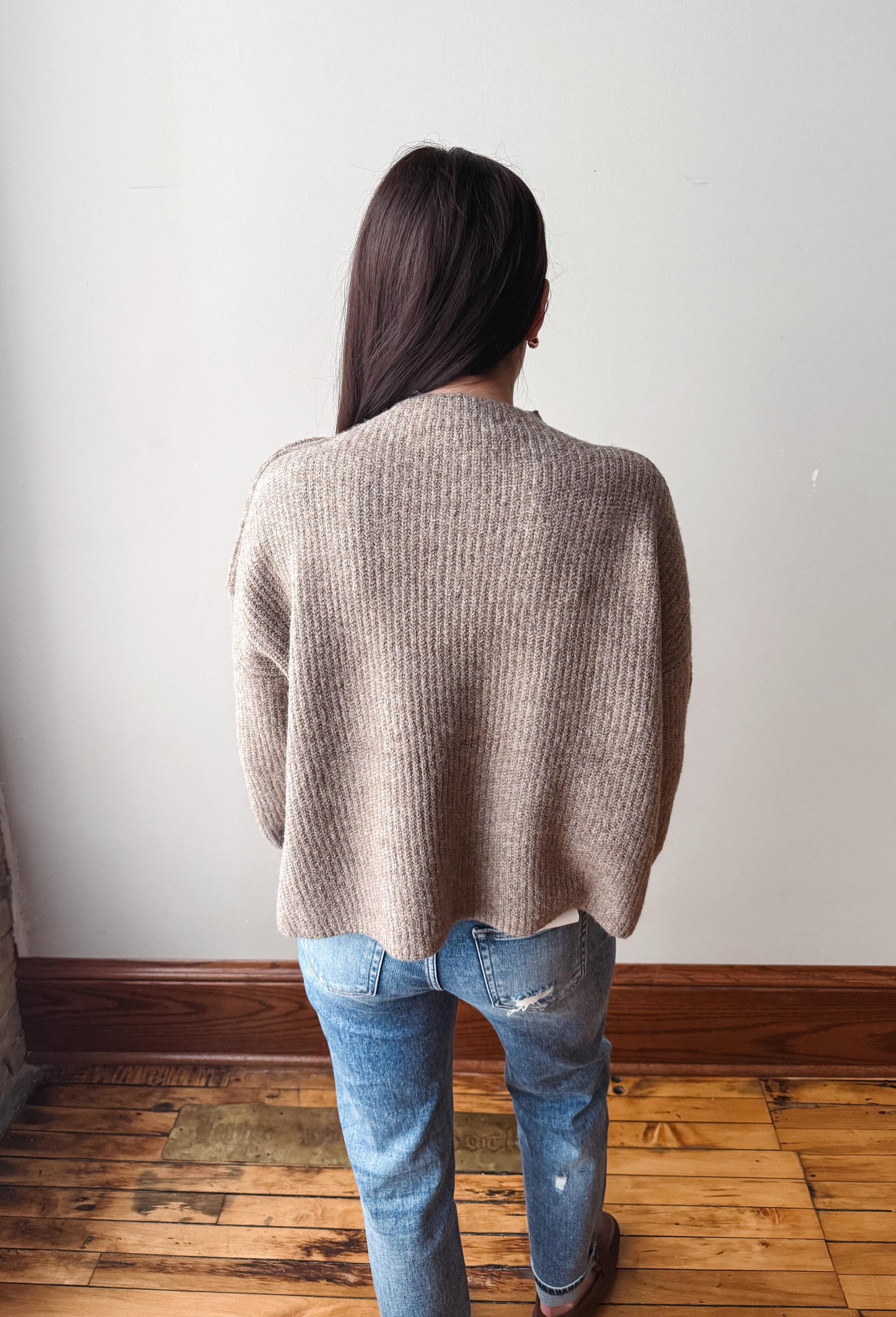 A woman viewed from behind wearing a beige ribbed knit sweater and blue jeans, standing on a wooden floor against a plain white wall.