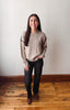 A woman stands in a room with a light gray wall and wooden floor, wearing a beige knit sweater with black shoulder detailing, black pants, and brown boots.