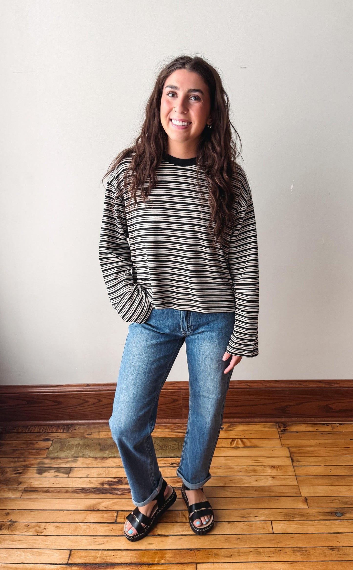 A woman wearing a black and white striped long-sleeve crewneck sweater, blue jeans, and black slide sandals, standing on a wooden floor against a plain off-white wall.