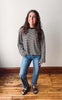 A woman wearing a black and white striped long-sleeve crewneck sweater, blue jeans, and black slide sandals, standing on a wooden floor against a plain off-white wall.