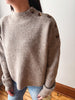 A person wearing a beige ribbed knit sweater with decorative brown buttons on the shoulders and upper sleeves, paired with blue jeans in an indoor setting.
