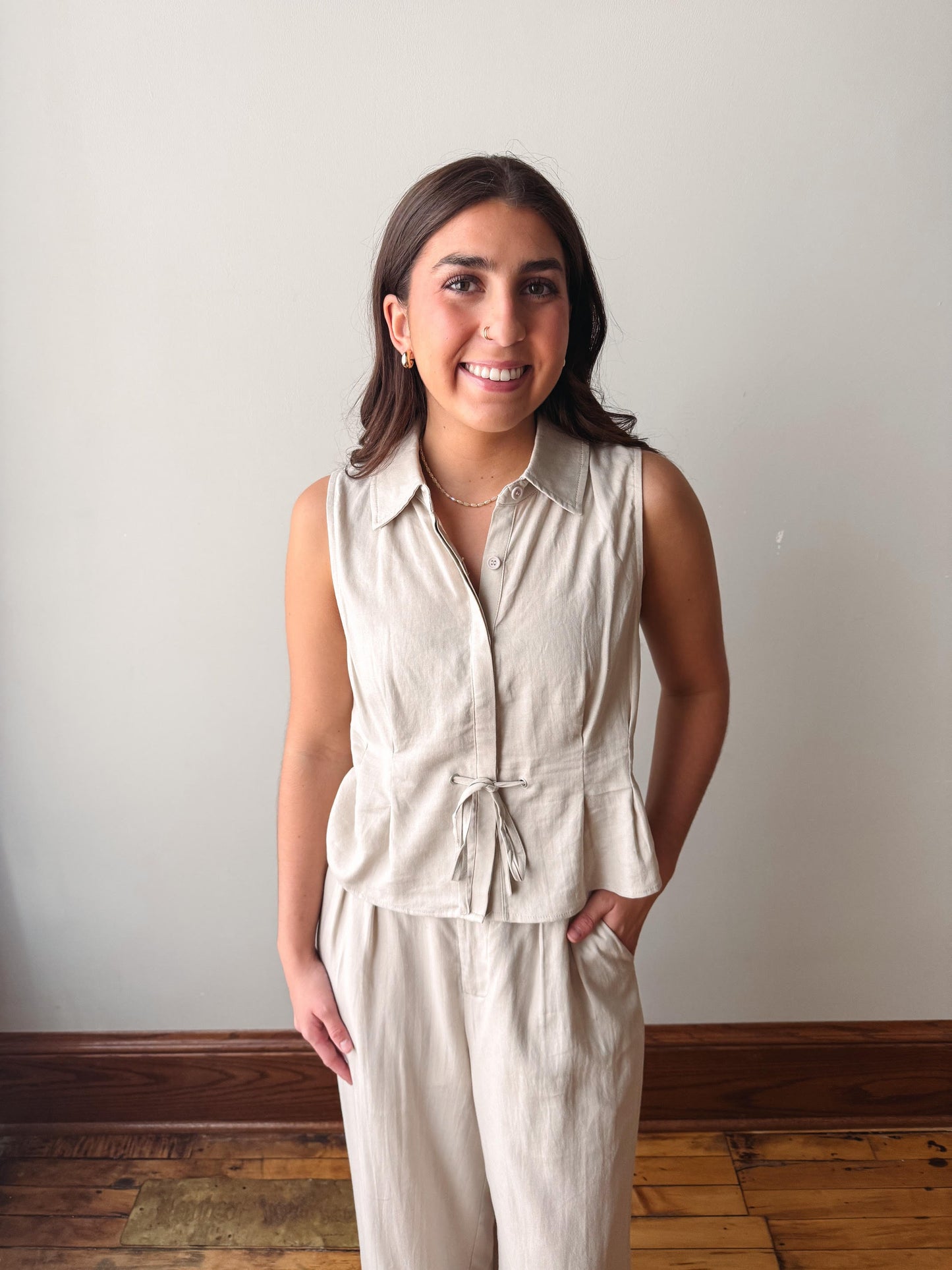 Woman wearing a beige sleeveless top and pants against a plain wall.