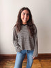 A smiling young woman with long brown hair stands indoors wearing a black and beige horizontally striped long-sleeve shirt with blue jeans, set against a plain off-white wall and wooden floor.