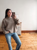 A woman takes a mirror selfie wearing a beige ribbed knit sweater and blue jeans in a room with light wood floors and a white wall.