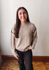 A woman with long dark hair stands indoors wearing a beige knit sweater with black decorative trim along the shoulders and sleeves, paired with black pants.