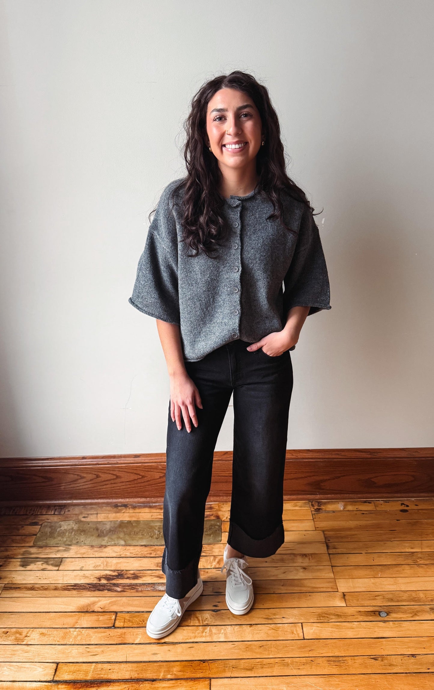 A woman stands on a wooden floor against a plain beige wall, wearing a gray short-sleeve button-front top, dark wide-leg jeans, and white sneakers.
