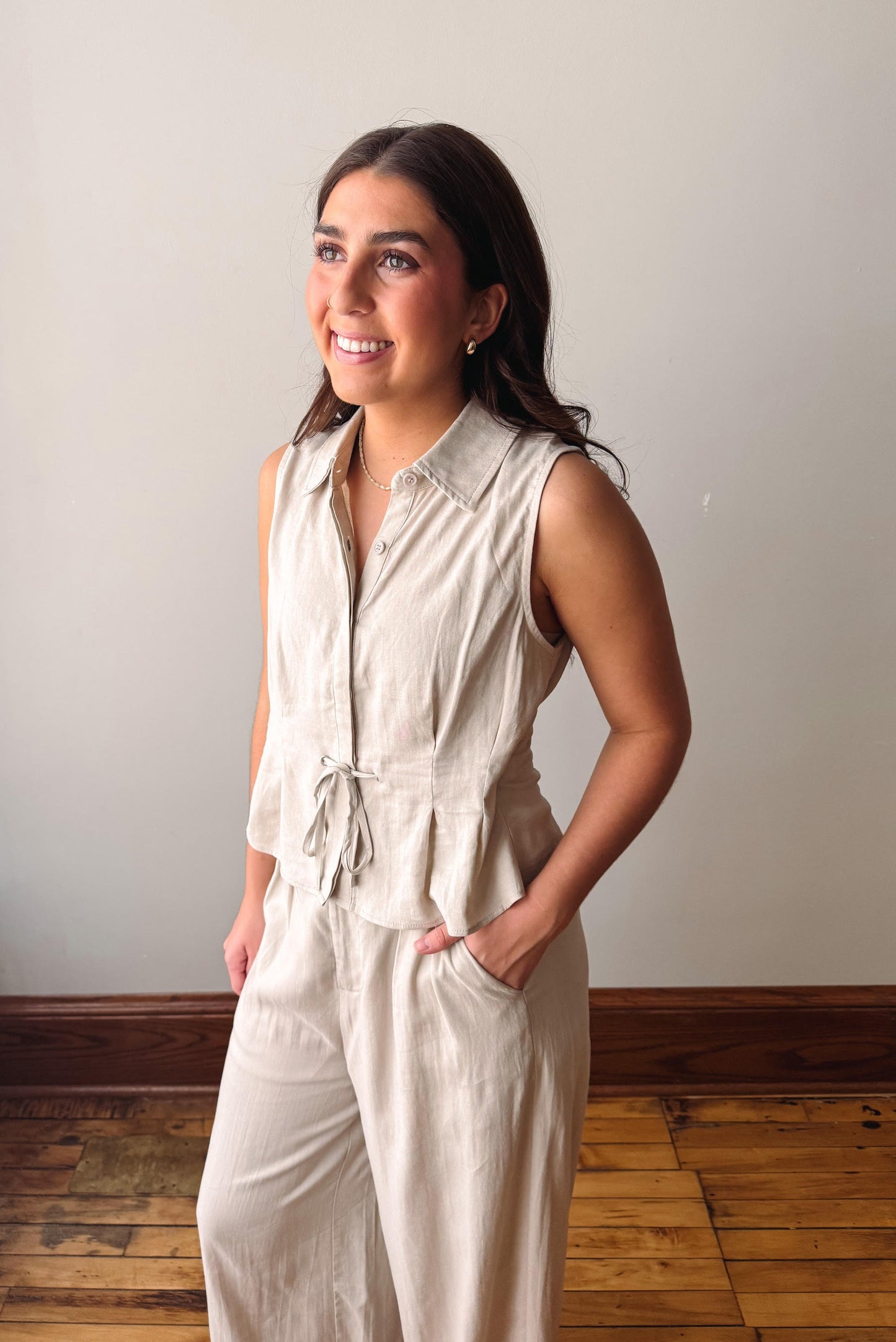 Woman wearing a beige sleeveless jumpsuit standing against a plain wall.