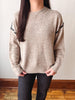 A person wearing a beige knit sweater with black decorative trim along the shoulders, paired with dark pants, standing indoors against a plain wall and wooden floor.