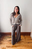 A woman stands in a room with a white wall and wooden floor, wearing a brown and cream striped knit cardigan with matching striped drawstrings over a brown wide-leg pants set.