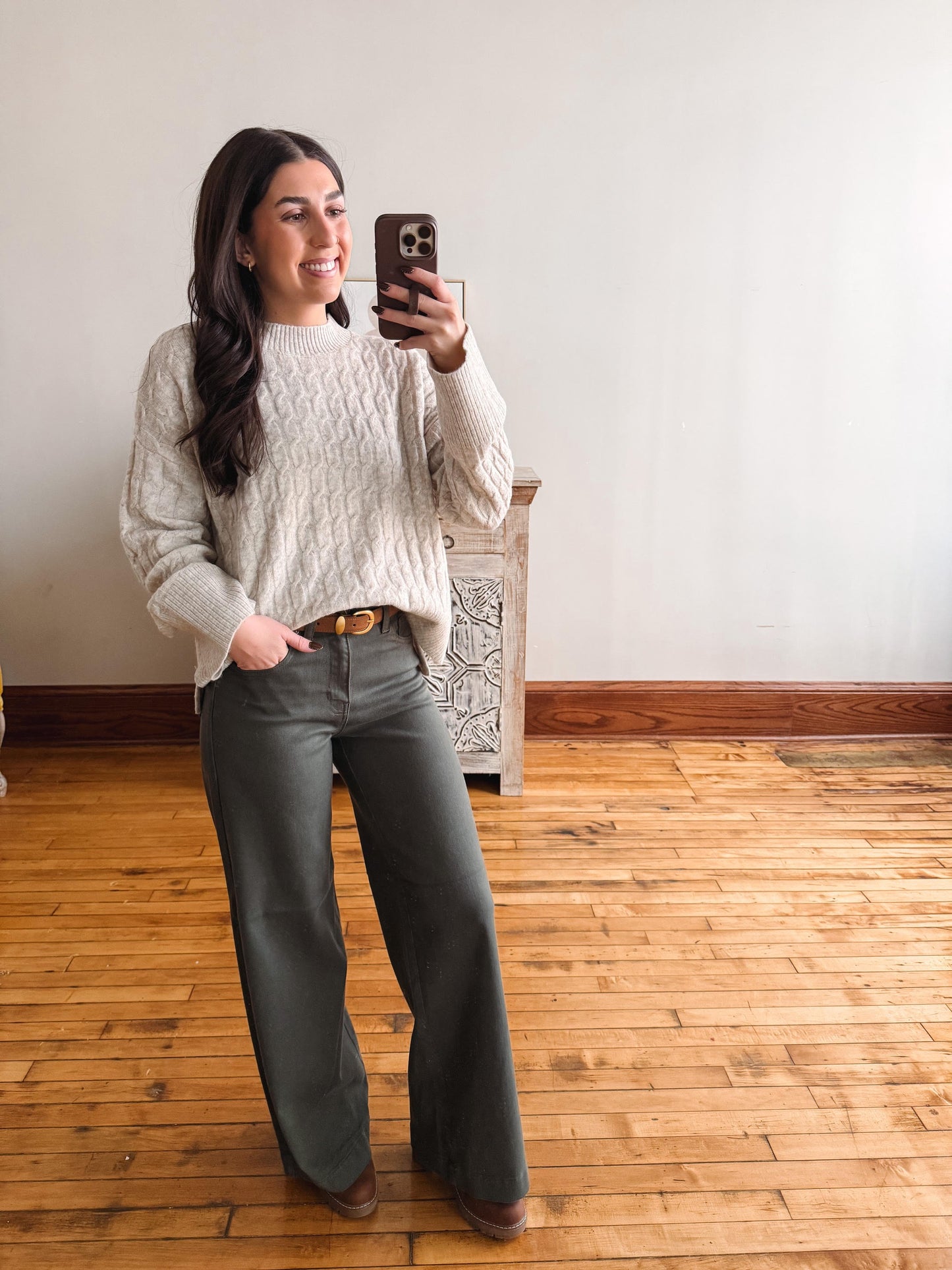 A woman takes a mirror selfie in a room with hardwood floors and a white wall, wearing a cream cable-knit sweater and dark olive green wide-leg trousers.