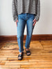 A person wearing blue straight-leg jeans with rolled cuffs, paired with a black and white striped long-sleeve top and black slide sandals, standing on a wooden floor against a plain wall.