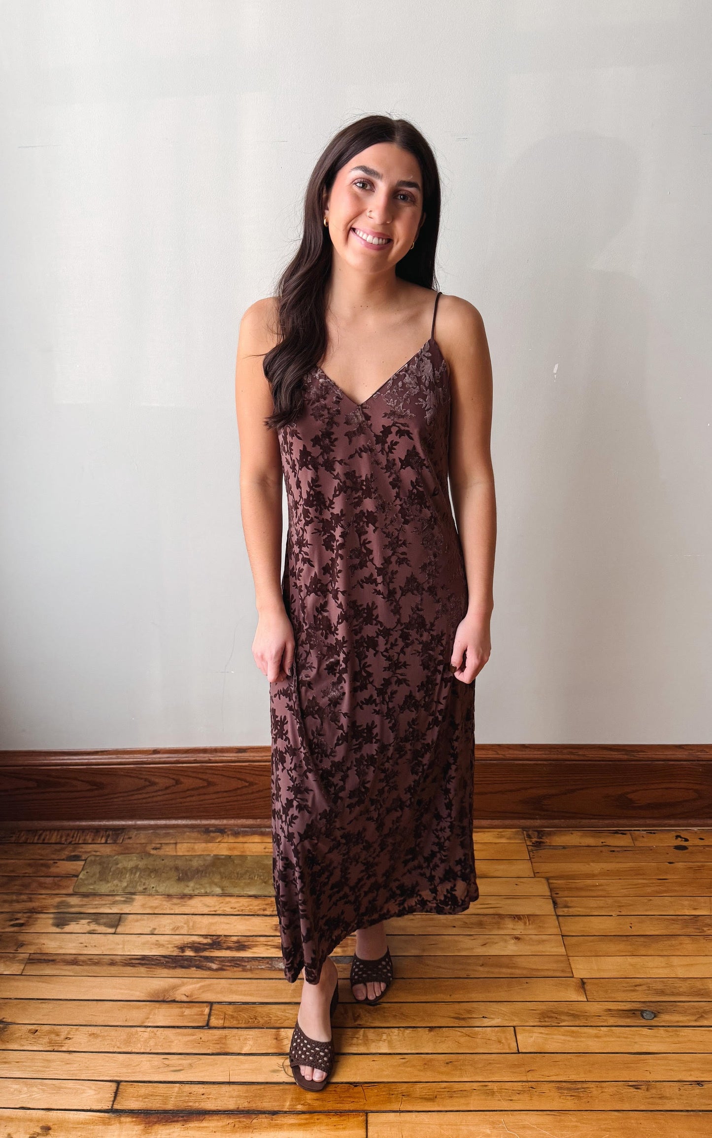 A woman modeling a dark brown floral lace slip dress with spaghetti straps, standing on a wooden floor against a plain off-white wall.