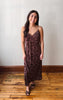 A woman modeling a dark brown floral lace slip dress with spaghetti straps, standing on a wooden floor against a plain off-white wall.