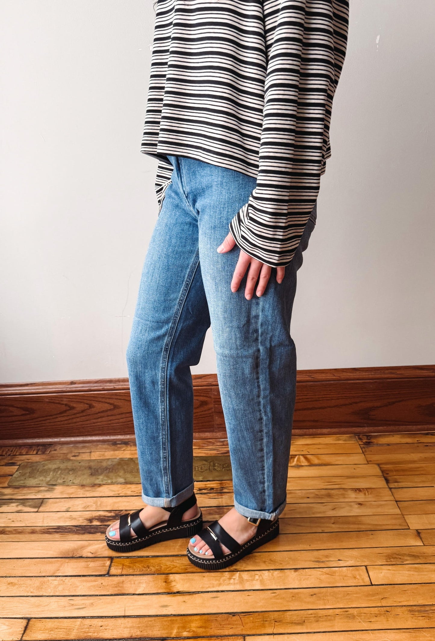 A person wearing light-wash blue jeans, a black and white striped long-sleeve shirt, and black sandals, standing on a wooden floor against a plain wall.