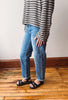 A person wearing light-wash blue jeans, a black and white striped long-sleeve shirt, and black sandals, standing on a wooden floor against a plain wall.