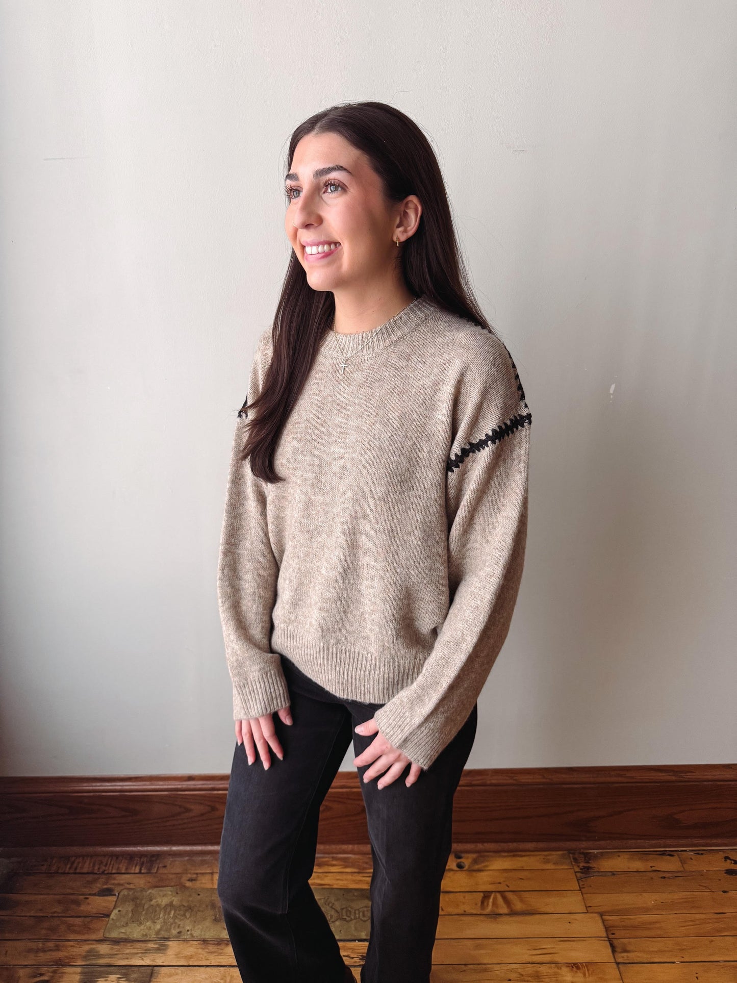 A woman wearing a light beige knit sweater with black zigzag trim on the shoulders, paired with black pants, standing in a room with a light gray wall and wooden floor.