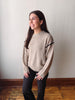 A woman wearing a light beige knit sweater with black zigzag trim on the shoulders, paired with black pants, standing in a room with a light gray wall and wooden floor.