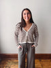 A woman stands indoors wearing a brown and white striped knit cardigan with a V-neck and front buttons, paired with loose taupe drawstring pants, against a plain light wall and wood floor.
