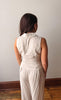 Woman wearing a beige sleeveless top and pants against a plain wall.