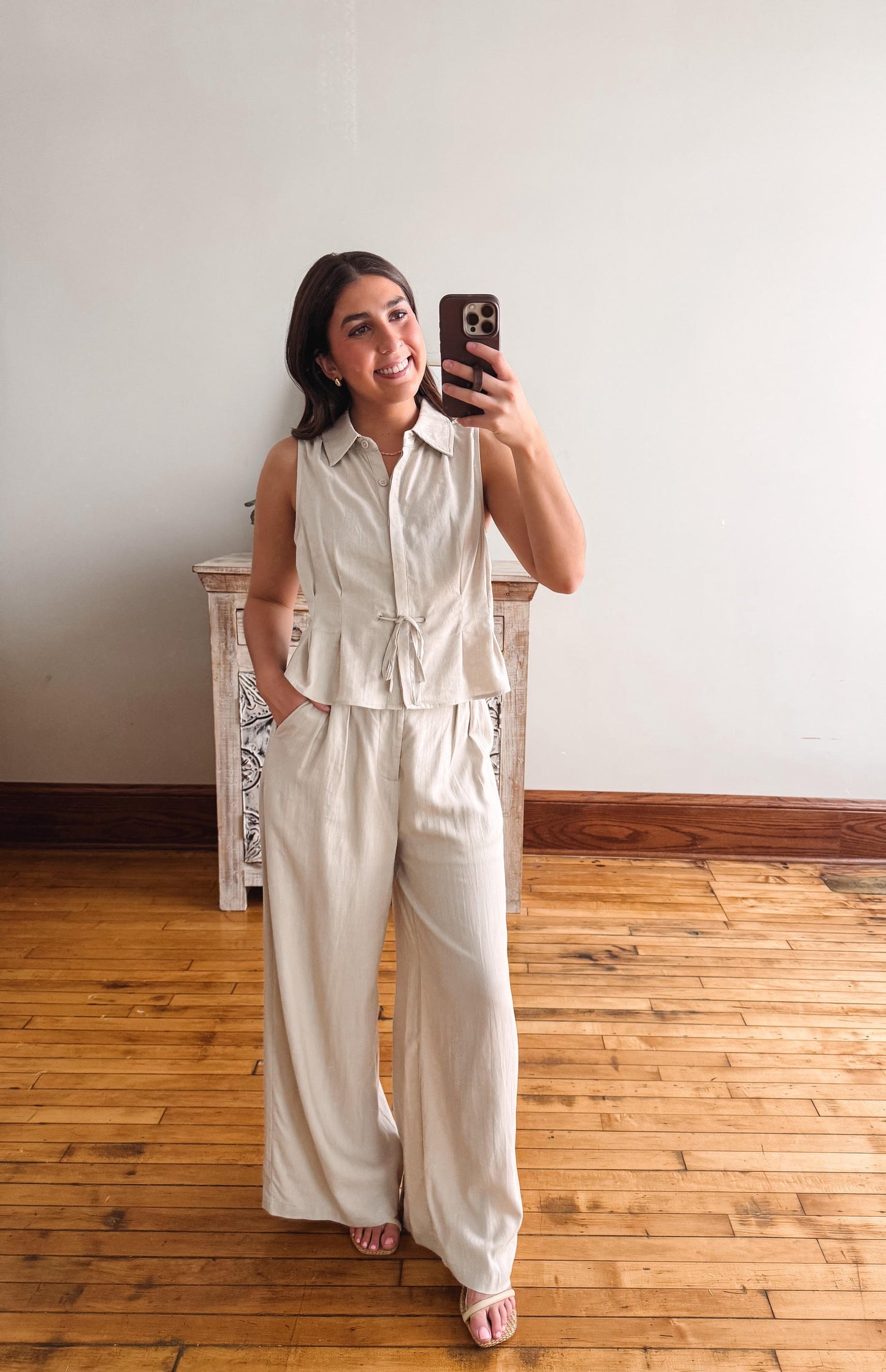 Woman taking a mirror selfie wearing a beige sleeveless top and wide-leg pants in a room with wooden flooring.