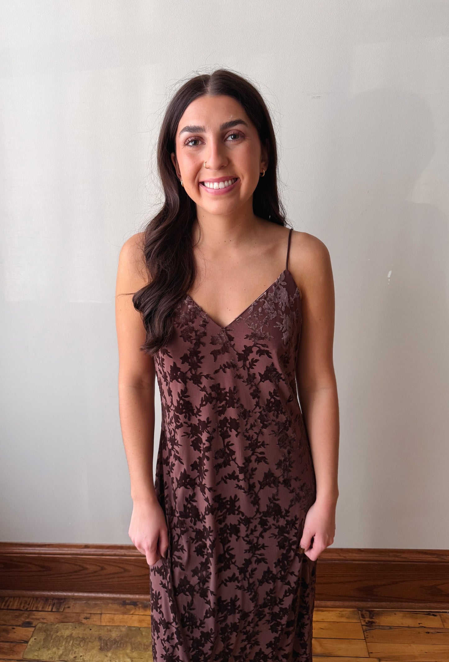 A woman with long dark hair stands indoors wearing a sleeveless, V-neck slip dress with a dark floral lace pattern over a muted brown base. She is posing in front of a plain off-white wall with a wooden baseboard and hardwood floor visible.