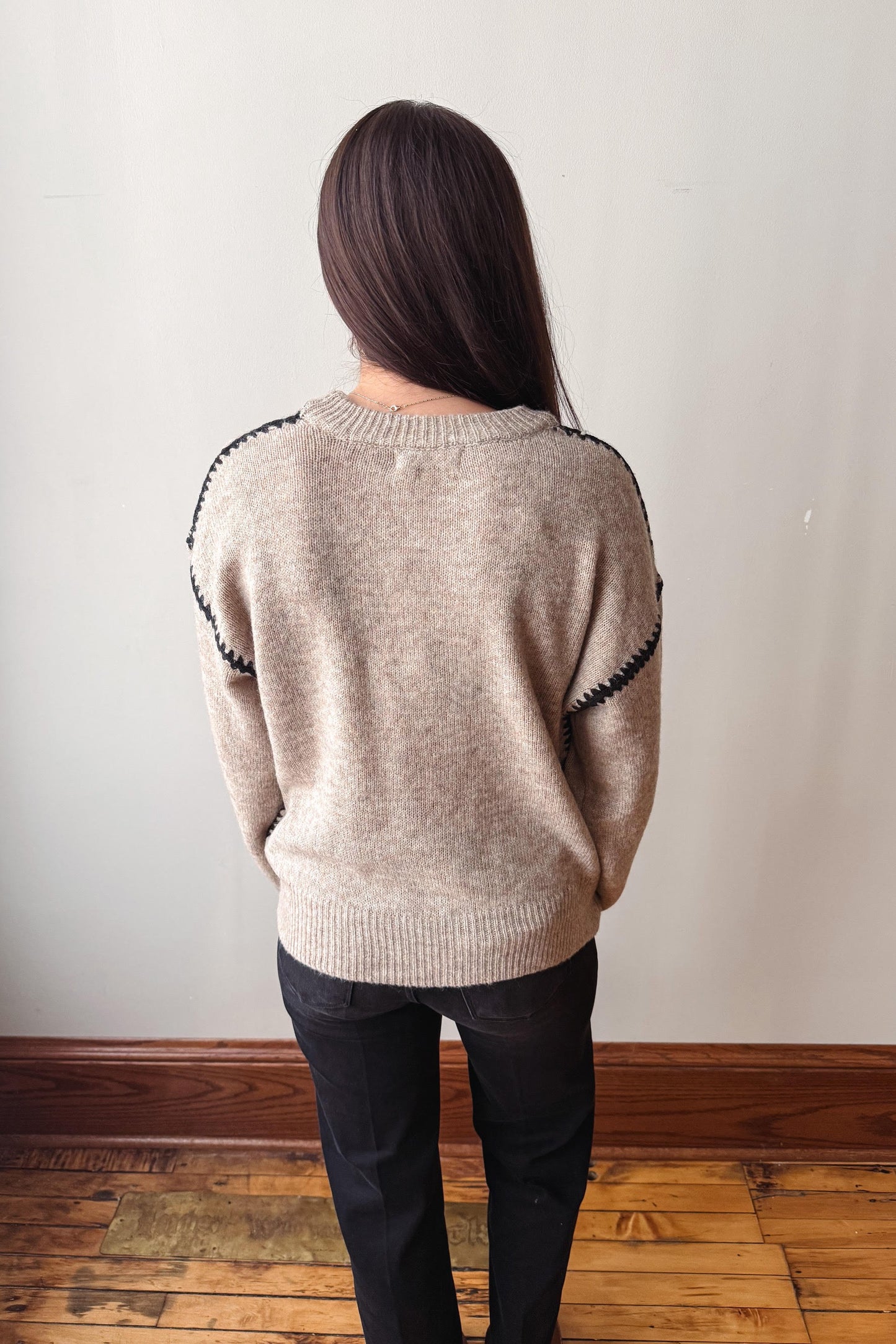 A woman viewed from behind wearing a light beige knit sweater with black decorative trim along the shoulders and dark pants, standing on a wooden floor against a plain off-white wall.