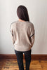 A woman viewed from behind wearing a light beige knit sweater with black decorative trim along the shoulders and dark pants, standing on a wooden floor against a plain off-white wall.