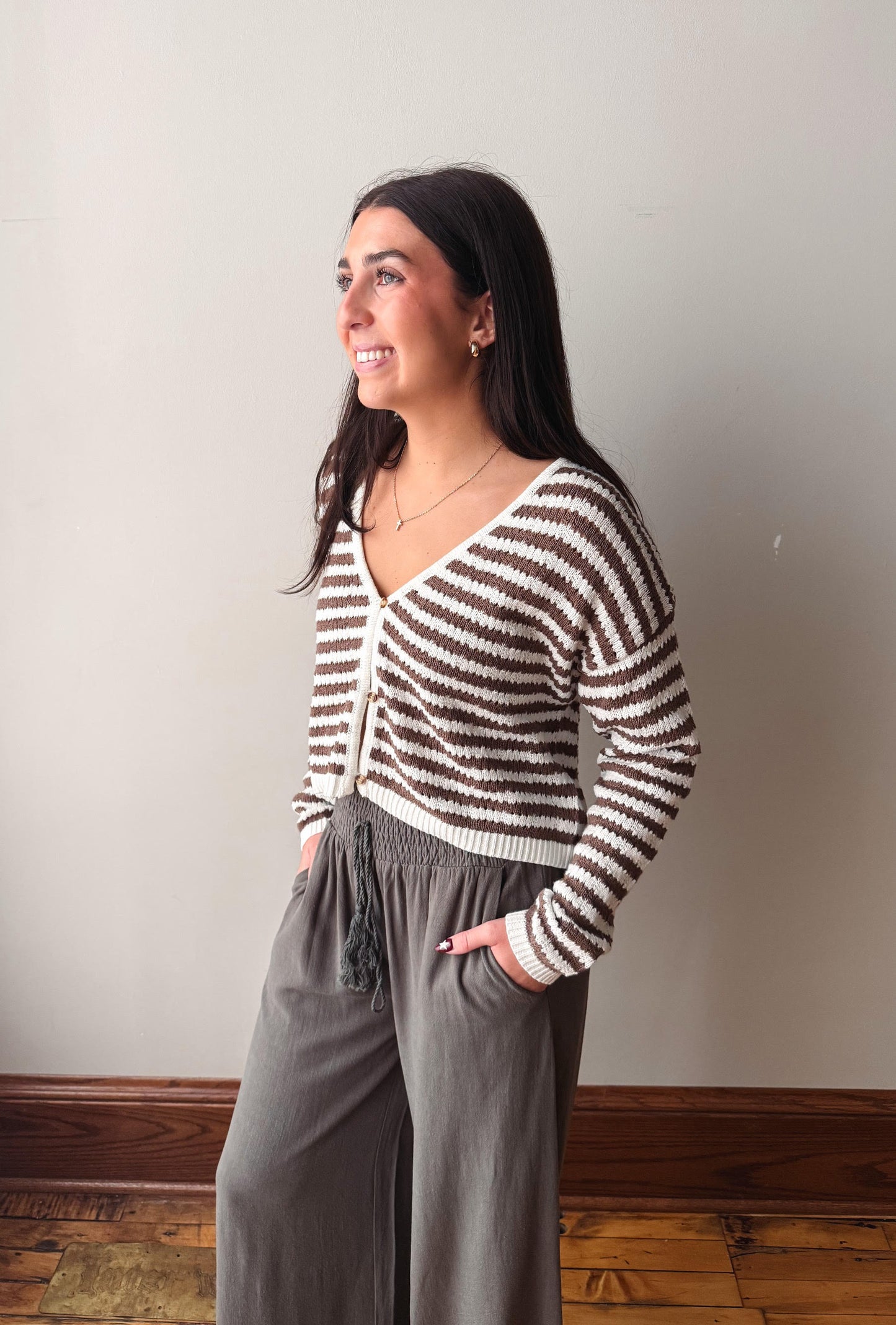 A woman wearing a brown and cream striped knit cardigan and gray wide-leg pants, standing in profile against a plain light gray wall with a wooden floor visible.