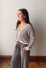 A woman wearing a brown and cream striped knit cardigan and gray wide-leg pants, standing in profile against a plain light gray wall with a wooden floor visible.