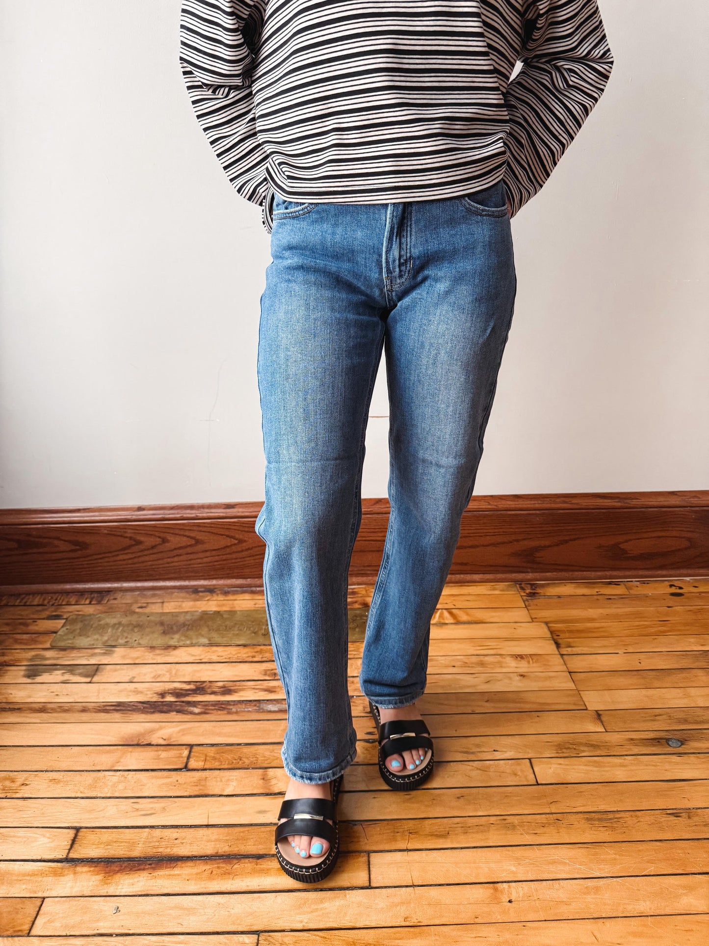 A person wearing medium-wash blue jeans, a black and white striped long-sleeve shirt, and black slide sandals, standing on a wooden floor against a white wall.