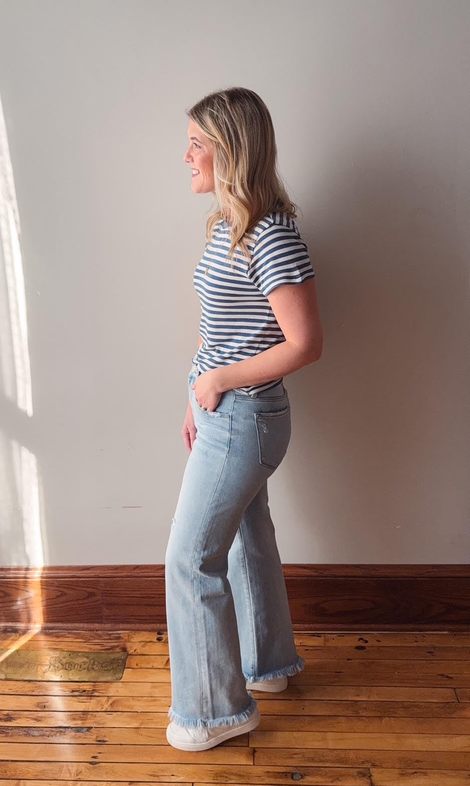 Woman wearing a striped shirt and light blue jeans standing against a white wall.