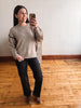 A woman takes a mirror selfie wearing a beige knit crewneck sweater with black trim on the sleeves, black pants, and brown ankle boots in a room with a light wall and wooden floor.