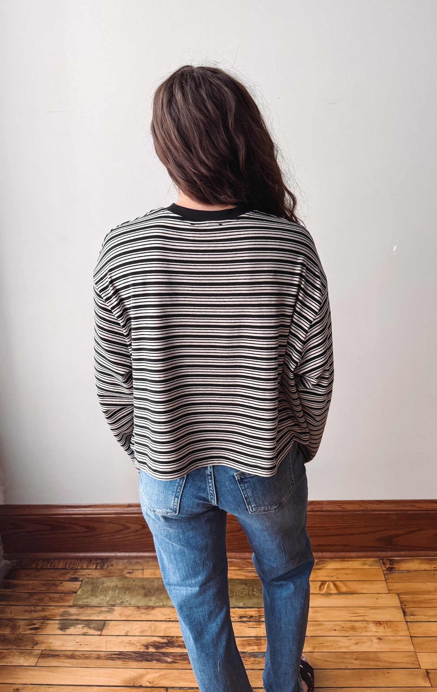 A person viewed from behind wearing a black and white horizontally striped long-sleeve shirt and blue jeans, standing on a wooden floor against a plain off-white wall.