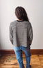 A person viewed from behind wearing a black and white horizontally striped long-sleeve shirt and blue jeans, standing on a wooden floor against a plain off-white wall.
