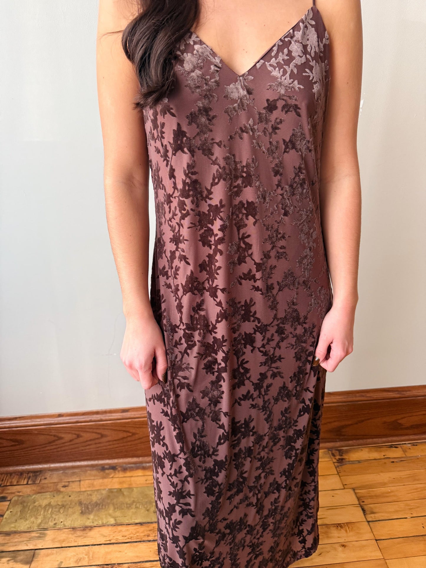 A woman wearing a sleeveless V-neck slip dress with a dark floral lace overlay on a dusty mauve-brown base, standing in a room with a light wall and wooden floor.