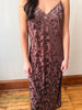 A woman wearing a sleeveless V-neck slip dress with a dark floral lace overlay on a dusty mauve-brown base, standing in a room with a light wall and wooden floor.