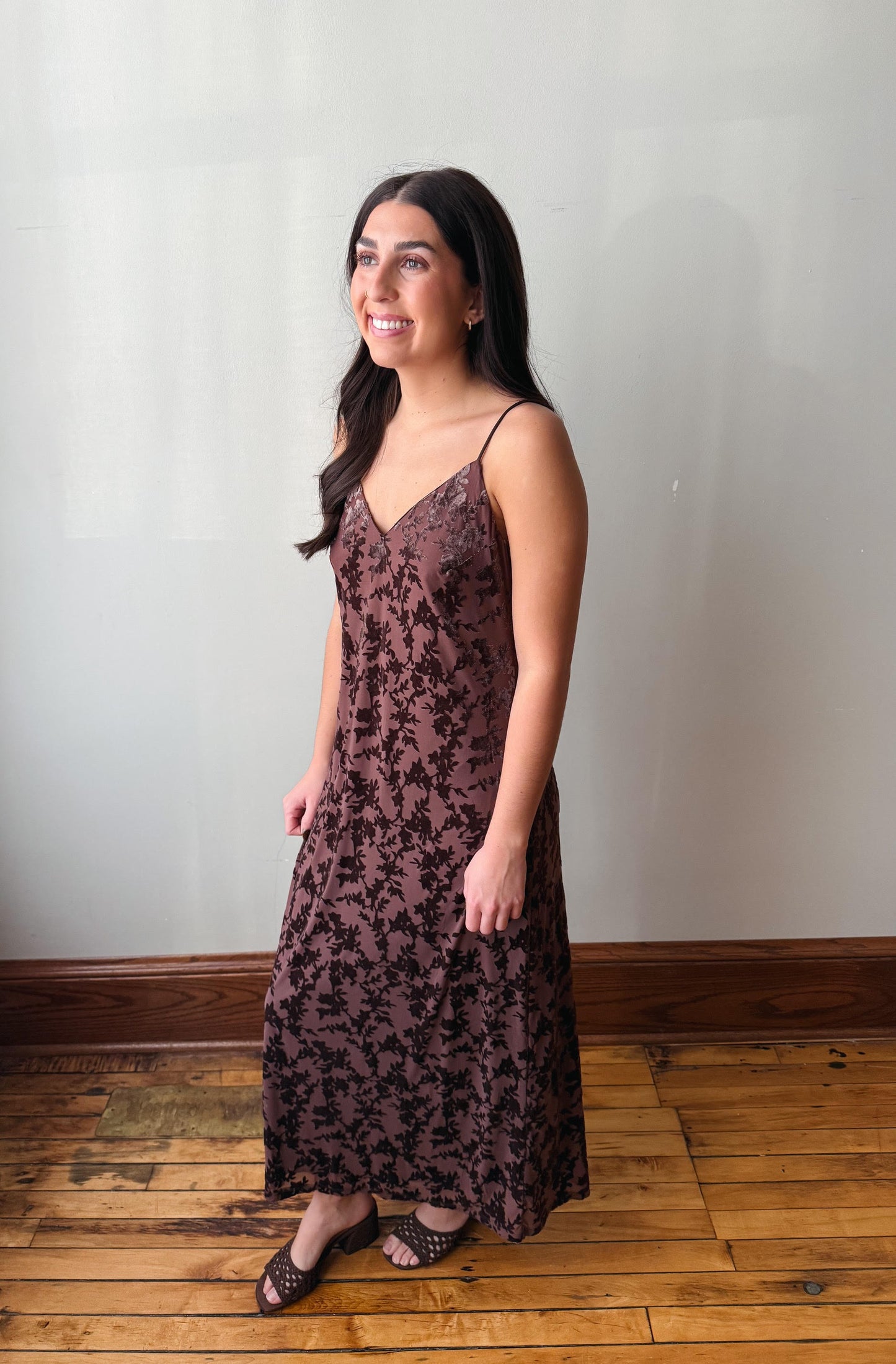 A woman modeling a sleeveless, floor-length slip dress with a dark floral pattern on a dusty rose-brown base, standing on a wooden floor against a plain light gray wall.