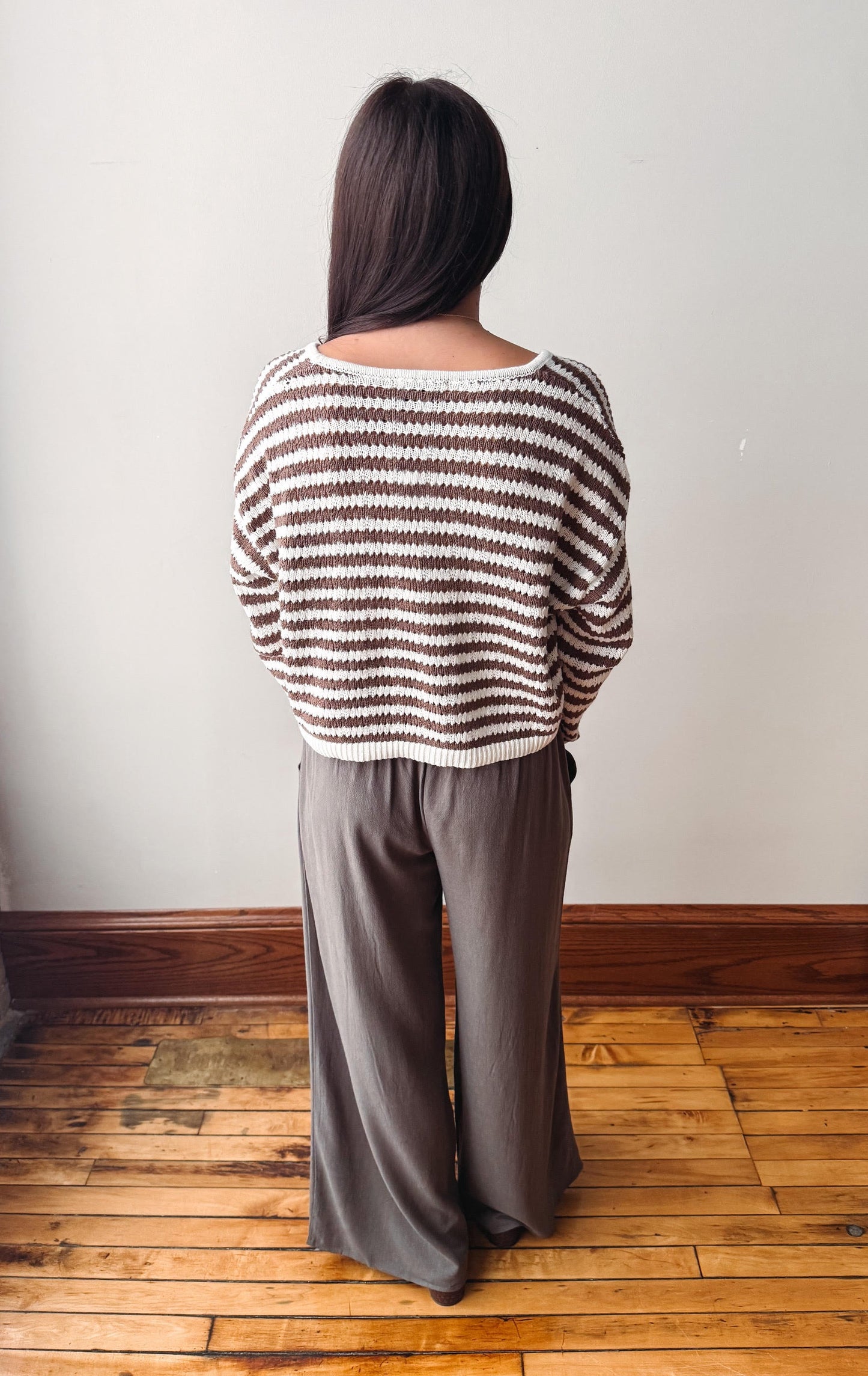 A woman viewed from behind wearing a brown and cream striped knit sweater and dark gray wide-leg pants, standing on a wooden floor against a plain wall.