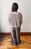 A woman viewed from behind wearing a brown and cream striped knit sweater and dark gray wide-leg pants, standing on a wooden floor against a plain wall.