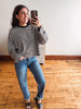 A woman takes a mirror selfie wearing a black and white horizontally striped long-sleeve shirt, blue jeans, and black slide sandals in a room with a light-colored wall and wooden floor.