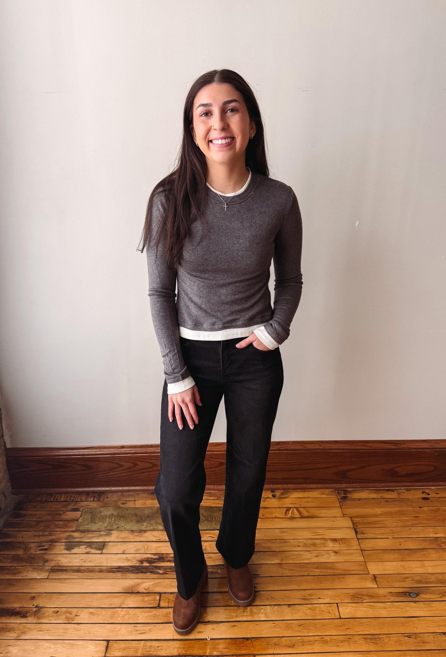 A woman stands in a room with a beige wall and wooden floor, wearing a dark gray long-sleeve knit top with white ribbed trim, black pants, and brown shoes.
