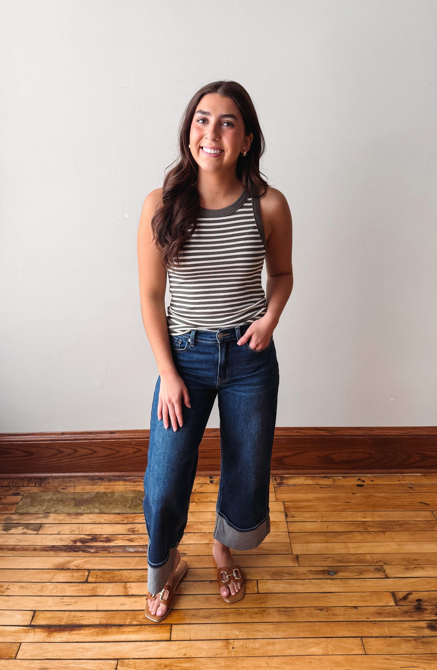 Woman wearing a striped tank top and jeans standing on a wooden floor with a white wall background